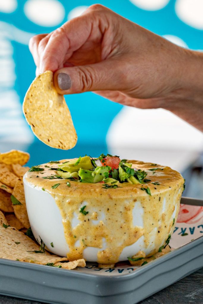 This is queso in a bowl with a hand to dip and tortilla chips in the background.